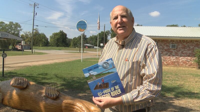 Daniel Brown stands with his book, Eli - Pride of the Yazoo River, next to a wooden carving of...